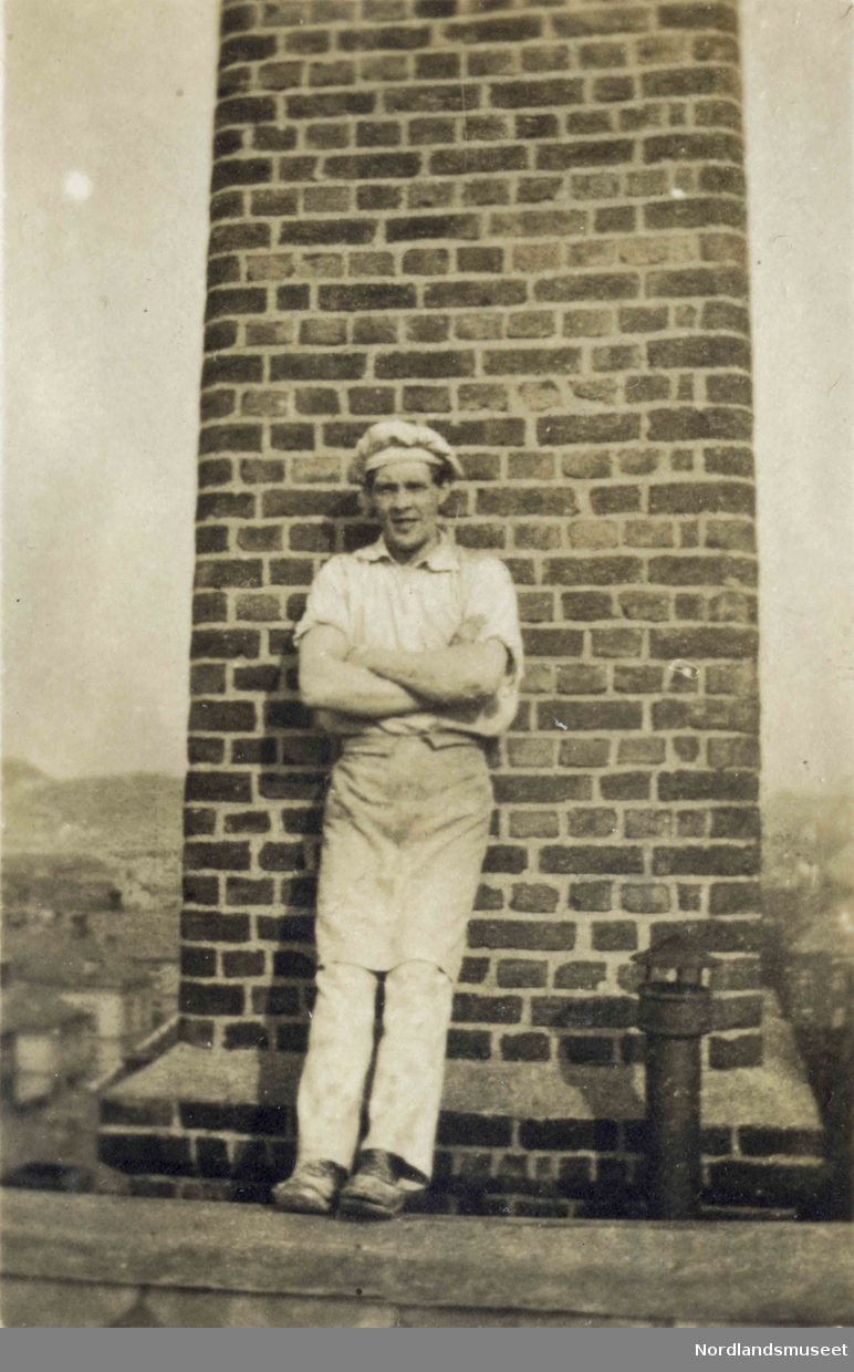 En mann i hvite klær står lent mot en murt pipe eller vegg.