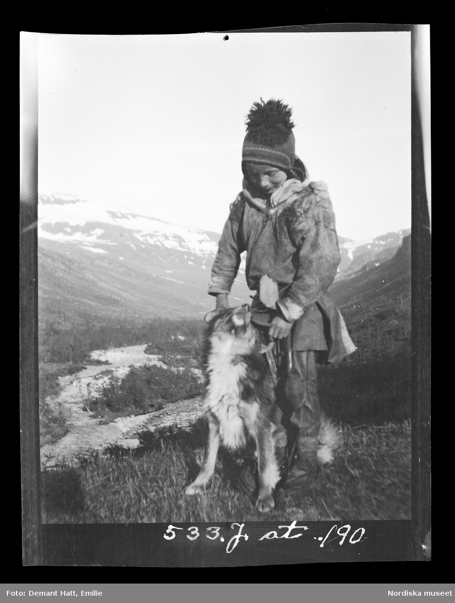 Ett barn leker med en hund (Curri), enligt bildtexten är pojken Andaras Ommas yngste son. I bakgrunden fjäll och en dalgång. Bilden ingår i en serie fotografier tagna av Emilie Demant Hatt i Sapmi mellan åren 1907 och 1916.