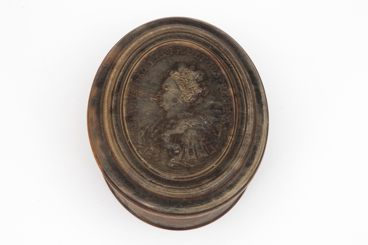 Ovalformet eske i horn (antagelig kuhorn) med løst lokk. Presset og dreiet med profilert lokkant omkring kronet dronningportrett i relieff. Portrettet er i profil vendt mot venstre og er antagelig av Anne, dronning av Storbritannia og Irland 1702-1714.