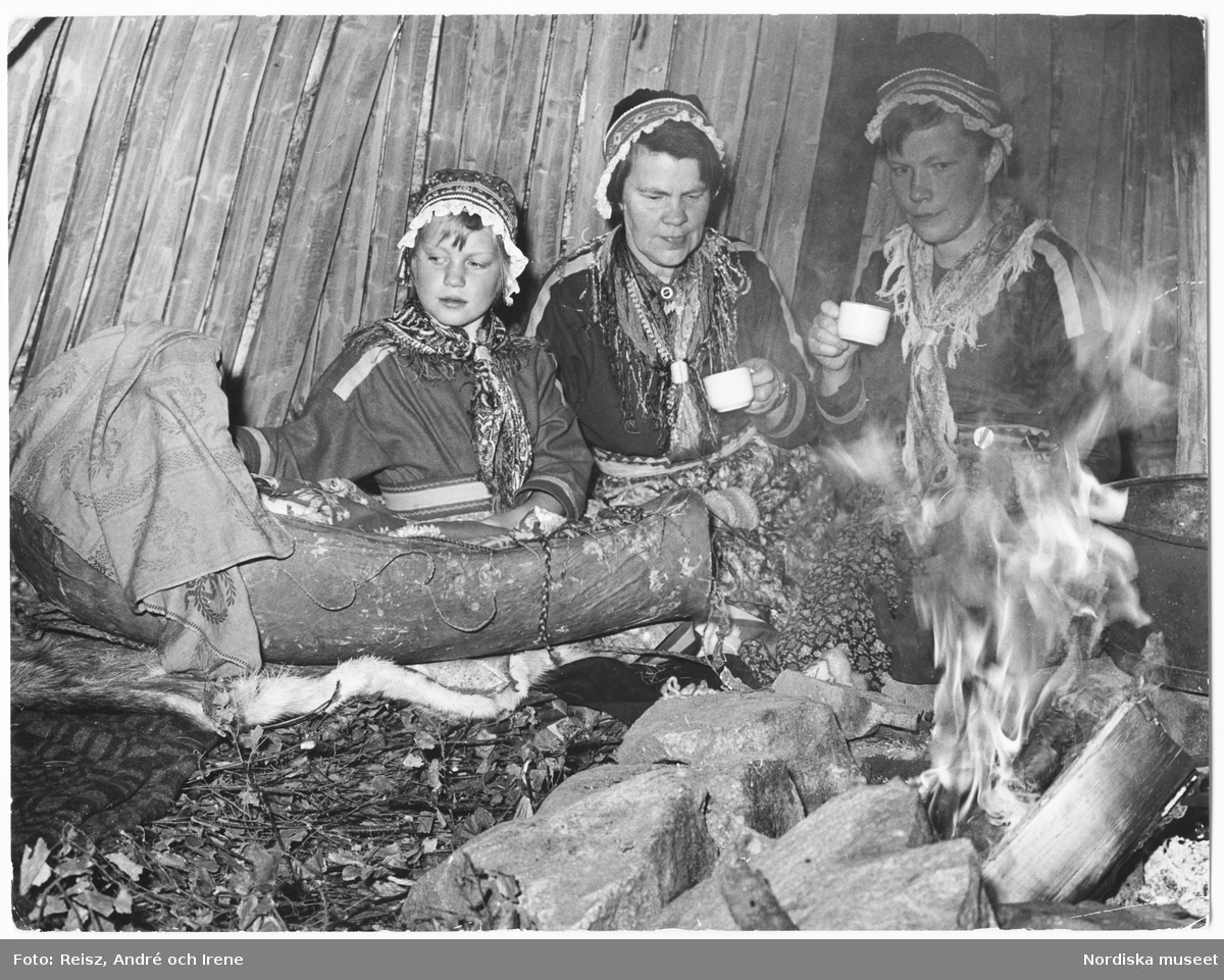 Samer, Norrland. Interiör av en lappkåta. Två kvinnor och en flicka, i samedräkter, sittandes med en samisk vagga vid den öppna elden.