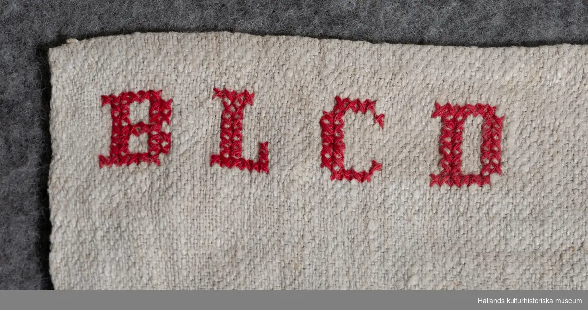 Linnelakan av grovt ofärgat (?) tyg. Märkt i korsstygn med rött bommulsgarn: "BLCD".