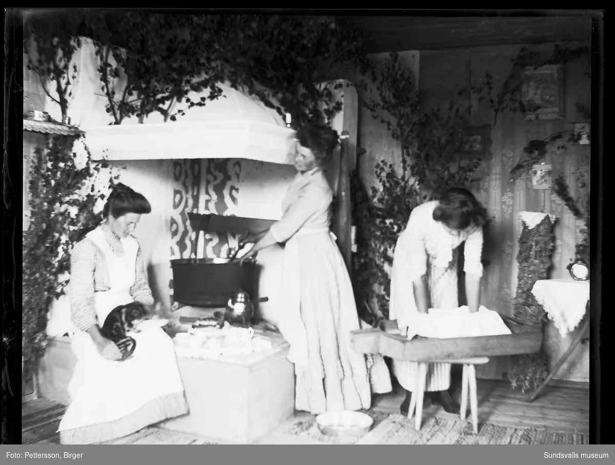 Fäbodbild från Klackbodarna i Stöde. Tre kvinnor i ett fäbodkök med öppen härd och björkris som pryder spiskåpan. Kvinnan längst till höger är sannolikt Emma Flemström, en av de andra kvinnorna har en katt i famnen.