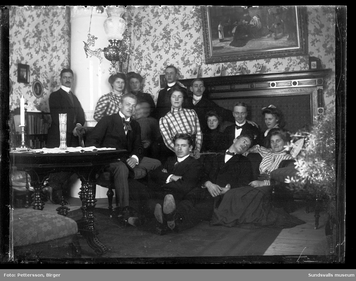 Gruppbild med yngre män och kvinnor inomhus i ett rum med kakelugn. Samma personer som SuM-foto035234.