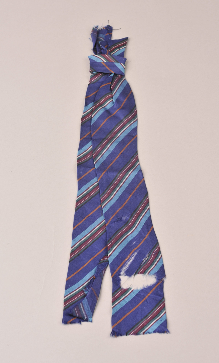 Del av slips i silke - sydd på skrå med striper i mørkeblått, turkis, grønt, hvitt, vinrødt, svart og lysebrunt. Knyttet øverst. Handsydd langs sidene - sårkant oppe og nede. 