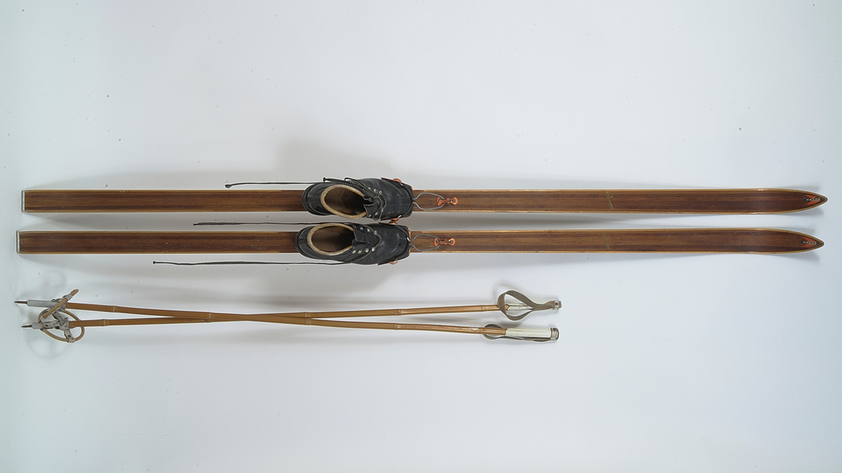 En av to skistaver, den andre A.06019-03.a. Selve staven er brun bambus. Håndtak, reimer og feste til trinse hvitt skinn. Metallpigg på enden. Trinsen av tre.