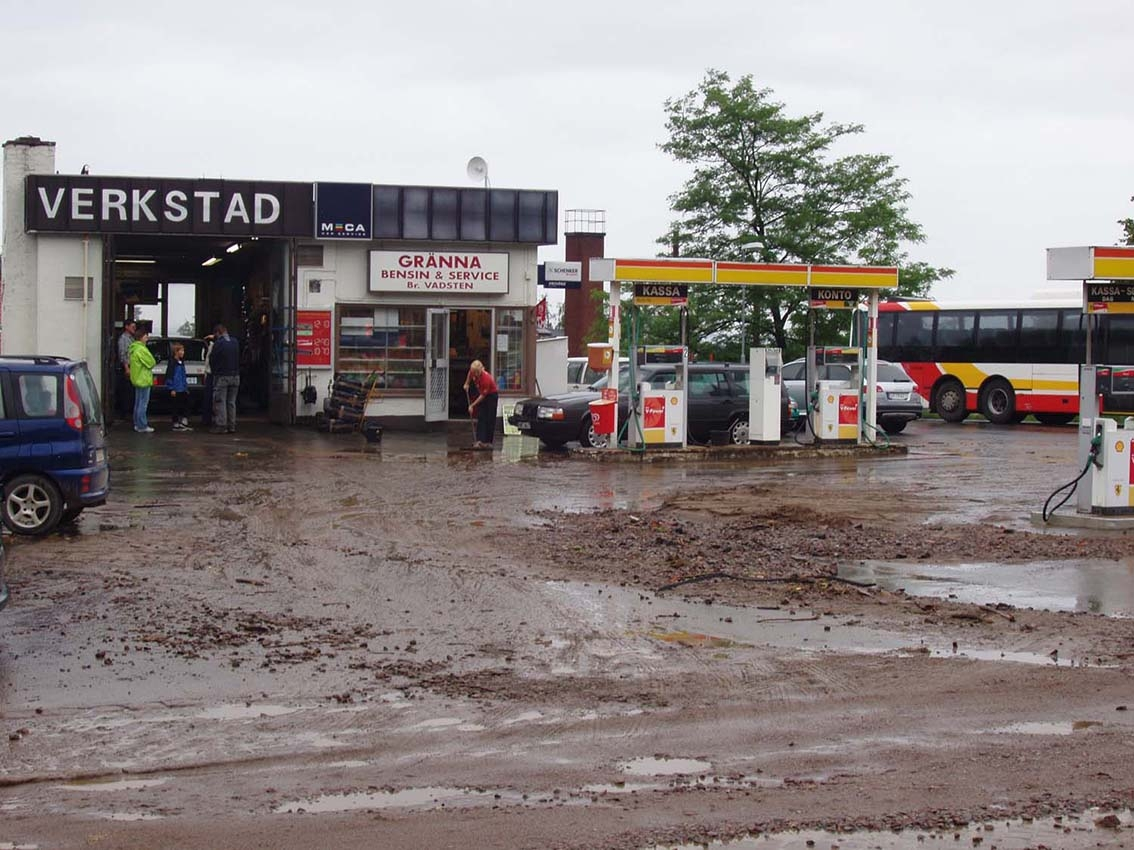 Grusmassor och vatten framför en bensinstation/verkstad i Gränna, en man står med kvast och borstar rent framför entén. En bil står i verkstaden.