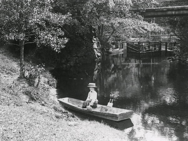 "Laxgård och hängbro i Uddaveka på 1920-talet." I förgrunden sitter en kvinna i stor hatt i en eka vid åstranden.