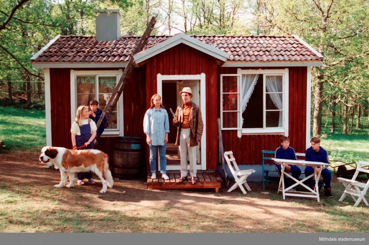 Sommarspelen/barnteaterföreställningen "Tjorven på Saltkråkan" 1996 vid Byxorna i Gunneboskogen. Troligtvis vid en repetition. Sex personer befinner sig vid en röd stuga. Från vänster står "Tjorven" med hunden "Båtsman" i koppel. Bakom henne står en pojke vid en stege. En kvinna och Per Andersson står vid entrén och till höger ses två pojkar som sitter i utemöbler. Omgivningen är fylld av grönska. Relaterade motiv: 2024_0525 - 0533.