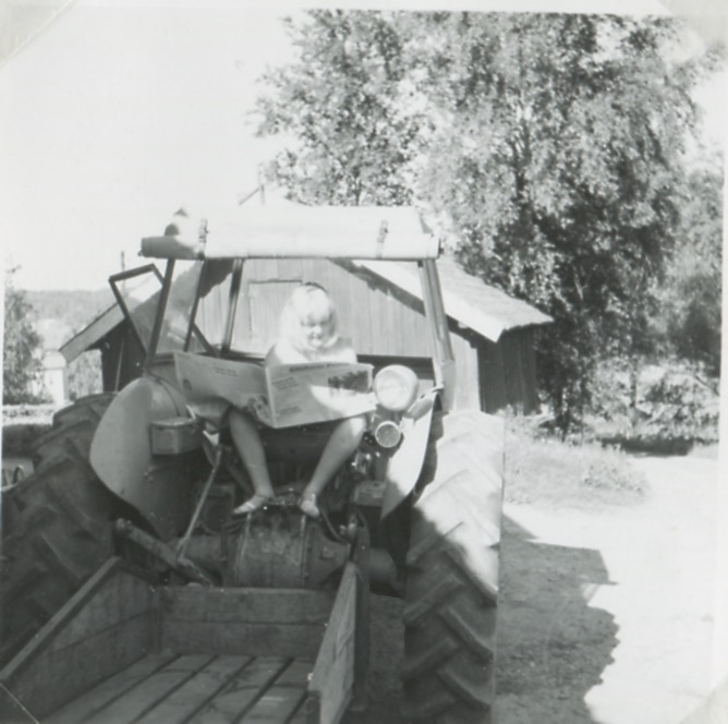 Ett barn sitter baktill på en traktor och läser tidningen, Backen eller Högen (nedre gården) 1960-tal. I bakgrunden skymtas en ekonomibyggnad.