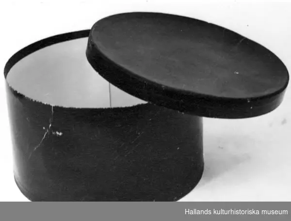 Hattaska av papp. Utvändig svart med moiréeffekt. Text på hattaskens undersida: "Gåva till Varbergs Museum av Fru Edgren Espenäs Ullared Tel 91134 ??".
