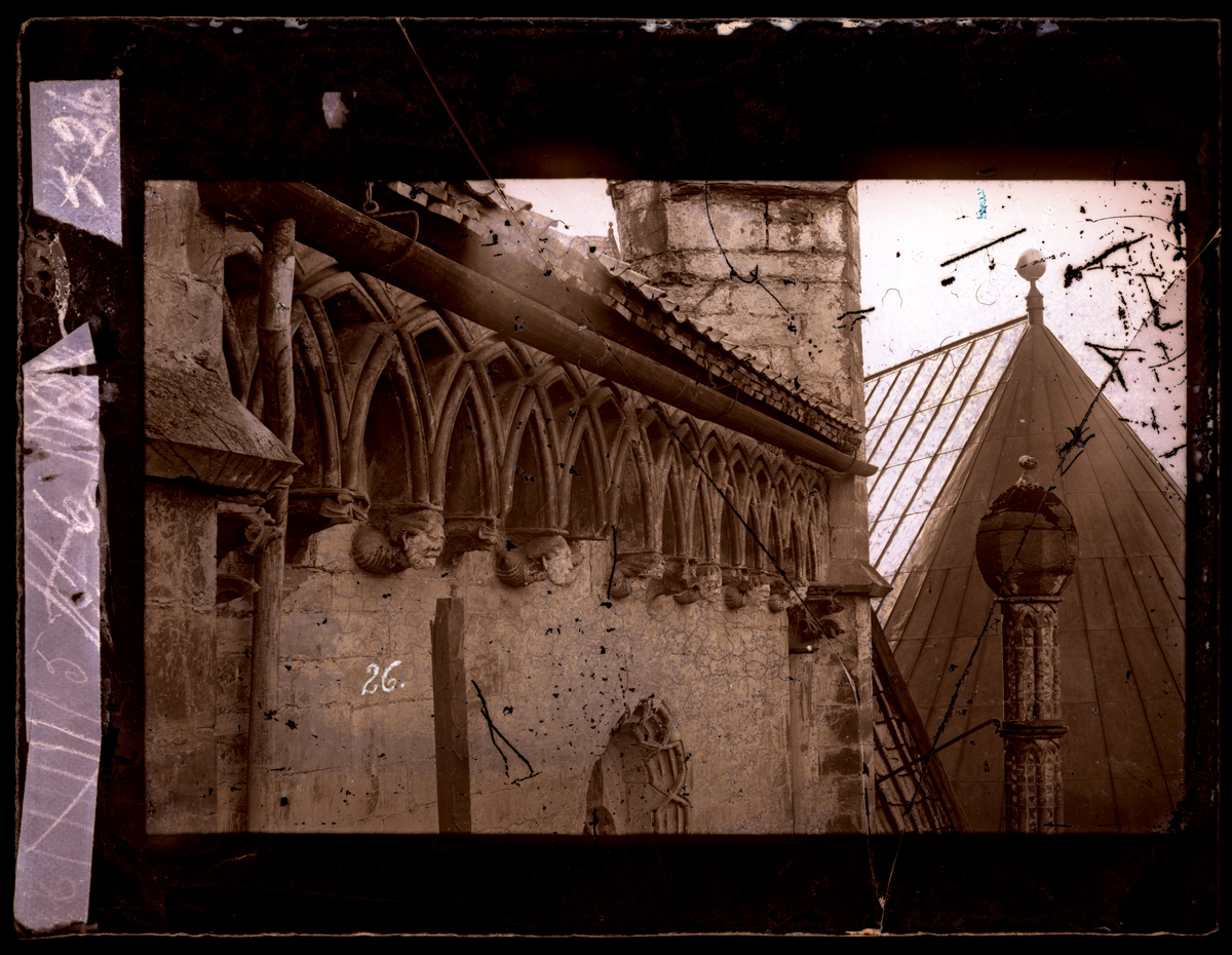 Øvre del av nord-østre vegg av oktogonen i Nidarosdomen. Buegesims med konsoller, flere i form av hodeskulpturer. Fial over nordre oktogonkapell til høyre i bildet. Takrenne av tre. Kapittelhusets tak i bakgrunnen (Kapittelhuset ble ferdig restaurert i 1871). Bildet er tatt før restaurering av oktogonen - et arbeid som pågikk i perioden 1872-1877.