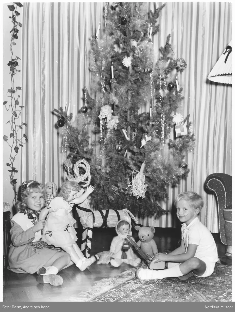 Två barn sitter på golvet vid julgranen. Flickan har en docka i knät, pojken håller i en leksakslyftkran.