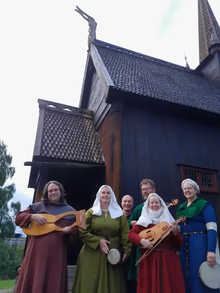 Middelalderensemble bestående av menn og damer i fargerike middelalderkostymer i rødt, grønt og blått. Damene har hvitt hodelin. De holder middelalderinstrumentene gitar, lyre, tamburin og tromme. De står foran en brun stavkirke.