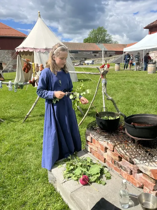 Ung jente i blå middelalderkjole som står foran et ildsted med gryter brukt til plantefarging