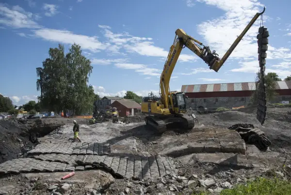 En gul gravemaskin med krokarm heiser på plass sprengningsmatter i gummi.