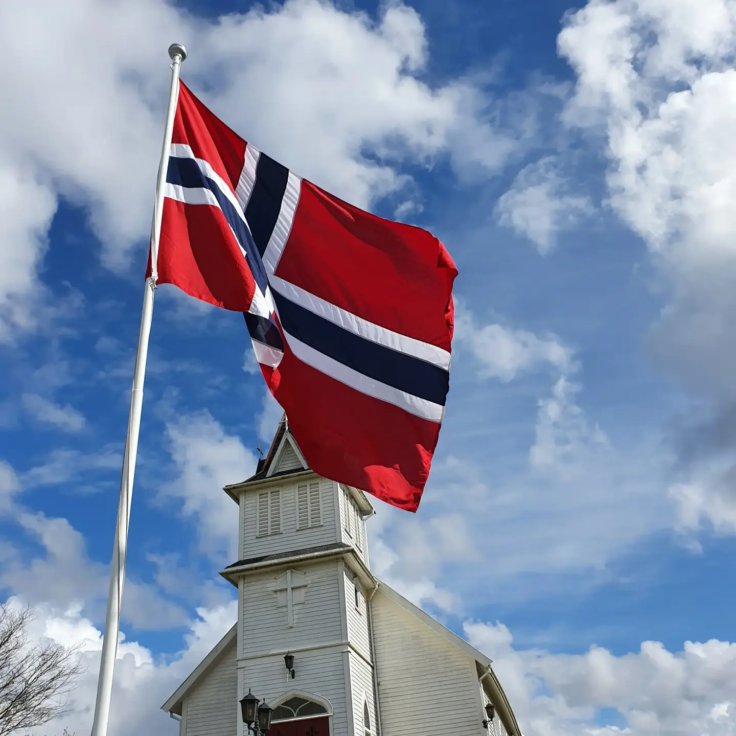 Norsk flagg vaien fremfor den hvite Emigrantkyrkja mot blå himmel med hvite skyer