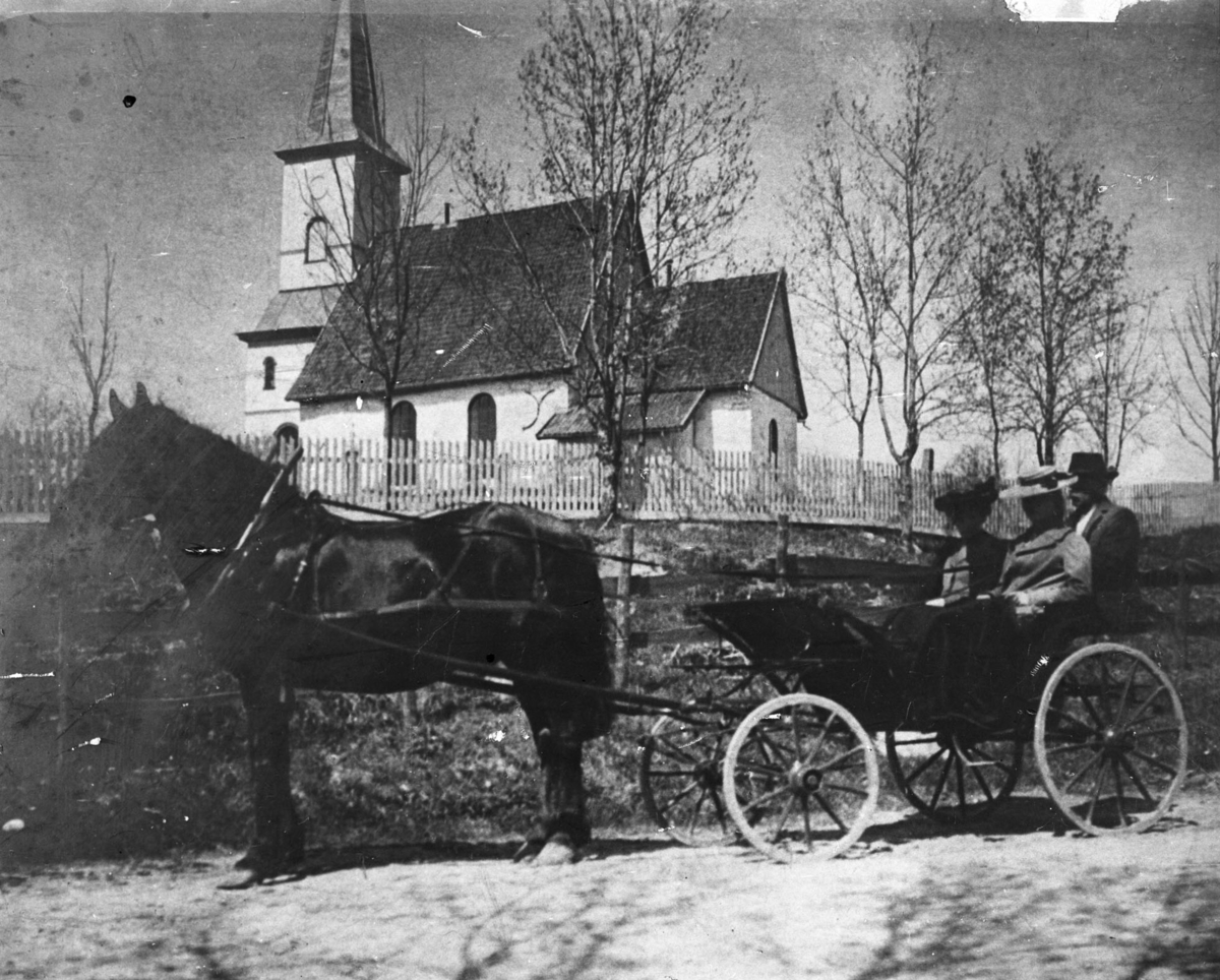 Hest og vogn med to kvinner og en mann utenfor Lørenskog kirke.
