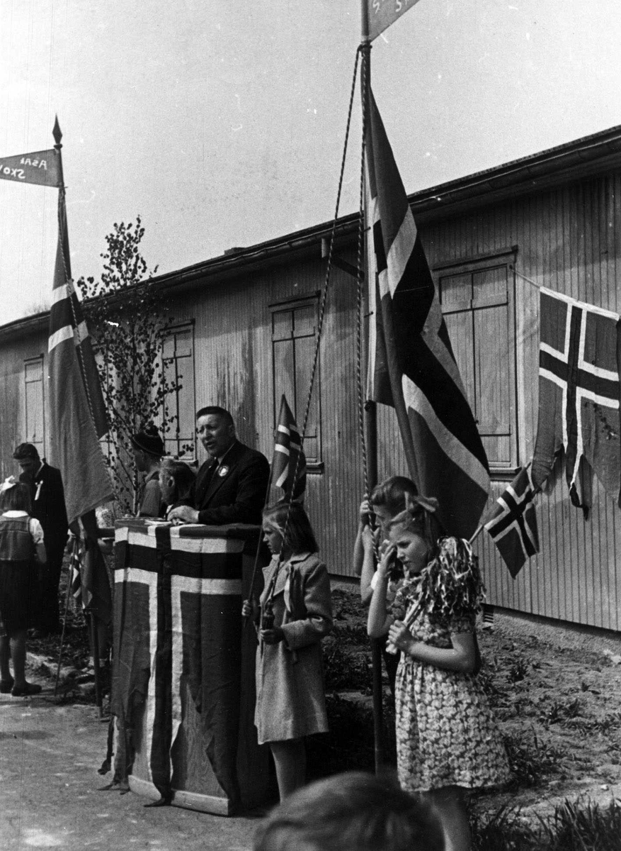 17. mai feiring på Kjeller 1945. Lærer Halvor Aasheim på talerstolen.
Norsk flagg p¨å talerstolen, to barn står flaggvakt. I bakgrunnen en brakke.