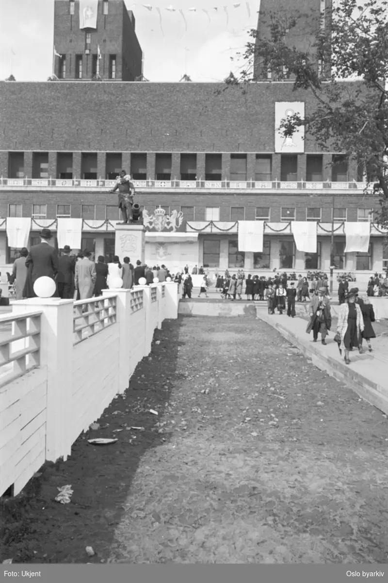 Festkledde mennesker på Rådhusplassen foran et smykket Oslo rådhus i anledning Kongens hjemkomst 7. juni 1945. Tordenskiold-statuen.