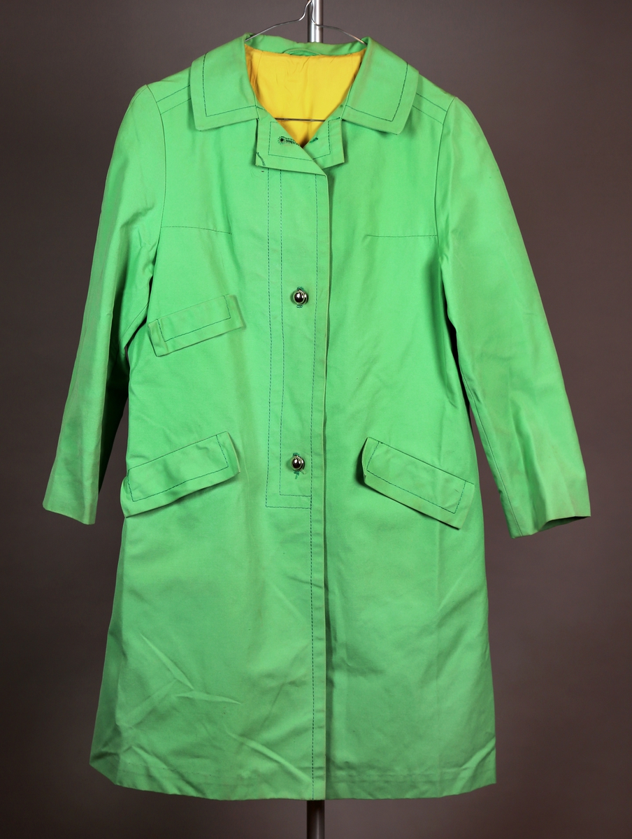 Grønn frakk med gult fôr, tre knapper i front av metall, og skråstilte lommer i front med klaffer over. 