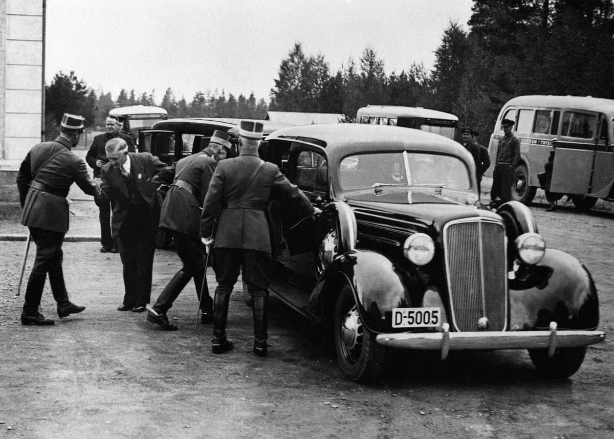 Elverum, Kong Haakon 7 og kronprins Olav i militæruniformer går inn i bilen til drosjeeier Vold, Chevrolet med registreringsnummer D-5005 årsmodell 1935 ved skolen i Strandbygdveien. Kronprins Olav hilser på drosjeeier Vold. General Sande til høyre.