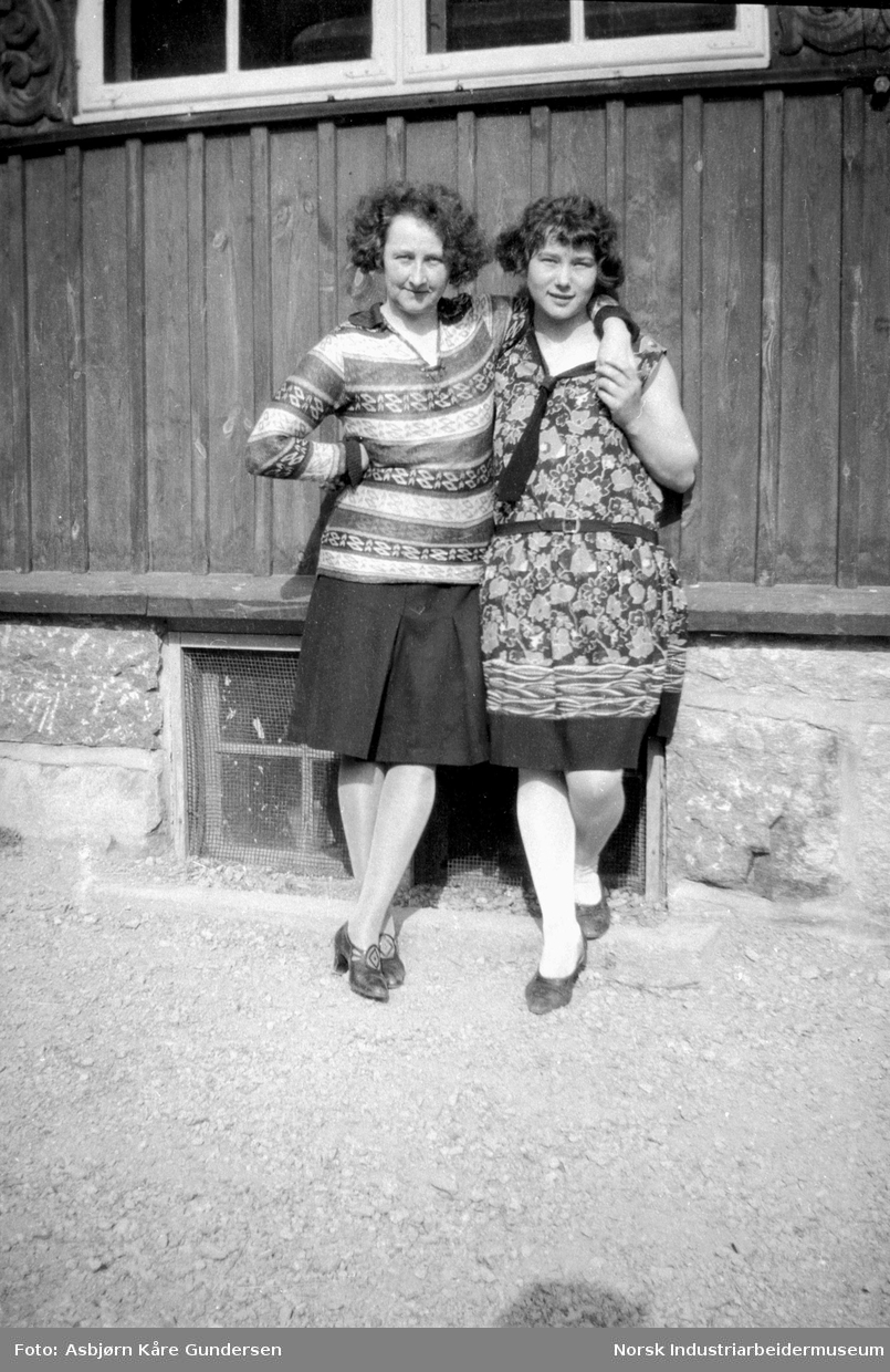 To kvinner i kameratslig positur foran en husvegg