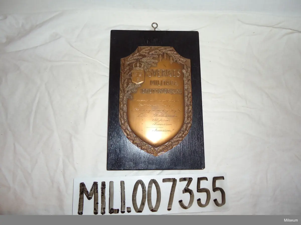 Prissköld av bronserad mässing, uppfästad på träplatta. "III. arméf.mil.idrottsdistr. Fälttävlan 17/7 1923.