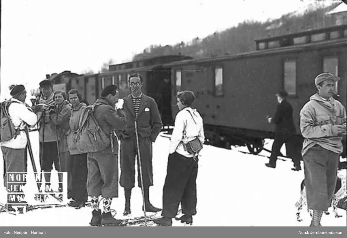 Skiturister på perrongen på Geilo stasjon, damplokomotiv med persontog i bakgrunnen