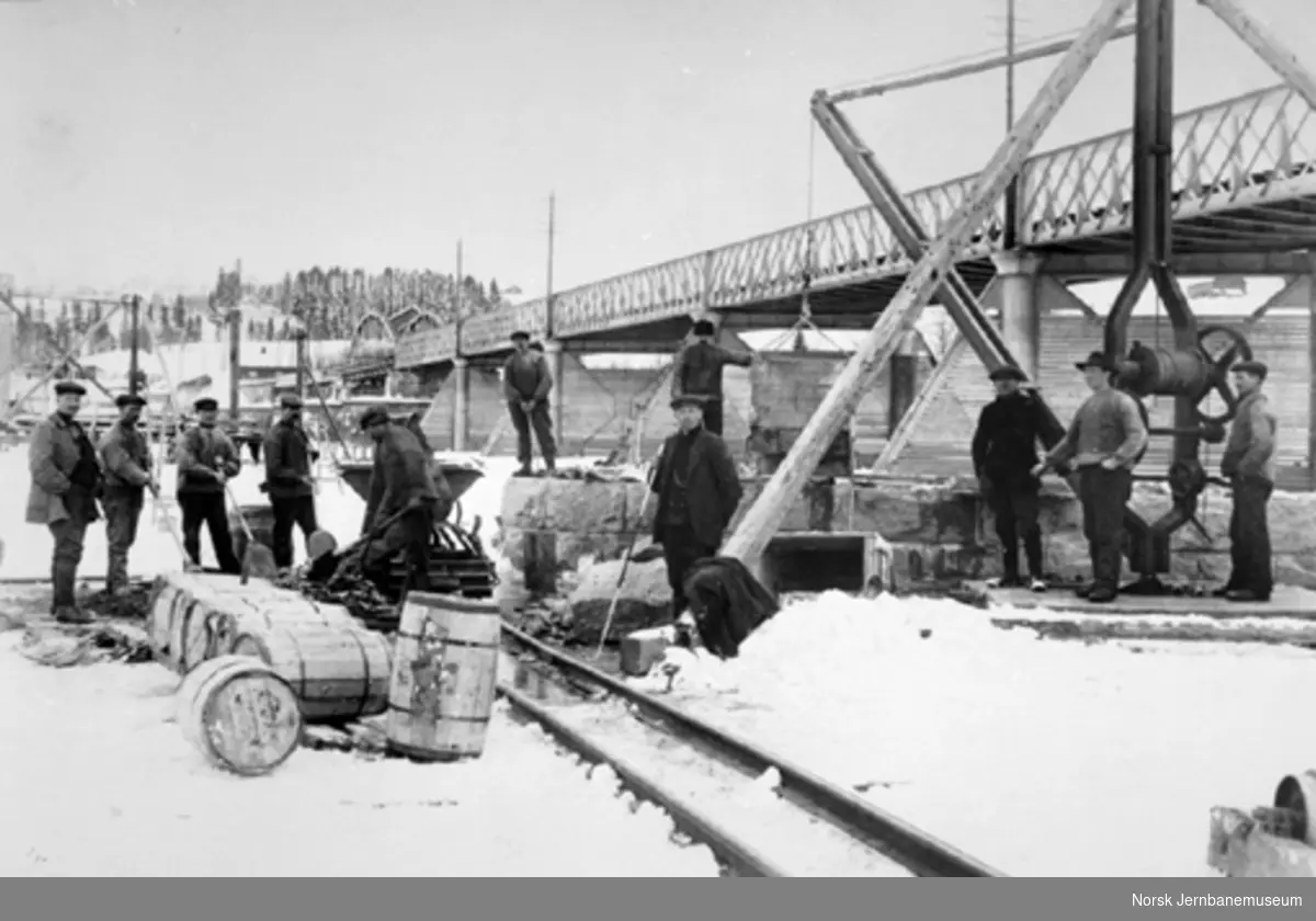 Arbeidslag på brygge (?) ved Fetsund bru ca. 1914. Vi ser bl.a. trallebane, kran og diverse tønner med sement. Til venstre oppsynsmann Ole Tohn.