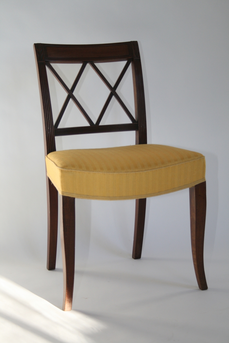 En av 8 like stoler i mahogni.
Ryggbrett med spiler i to diagonale kryss, og riller. Stoppet sete trukket med gult  stoff. Trekket er sekundært (1960-tall)