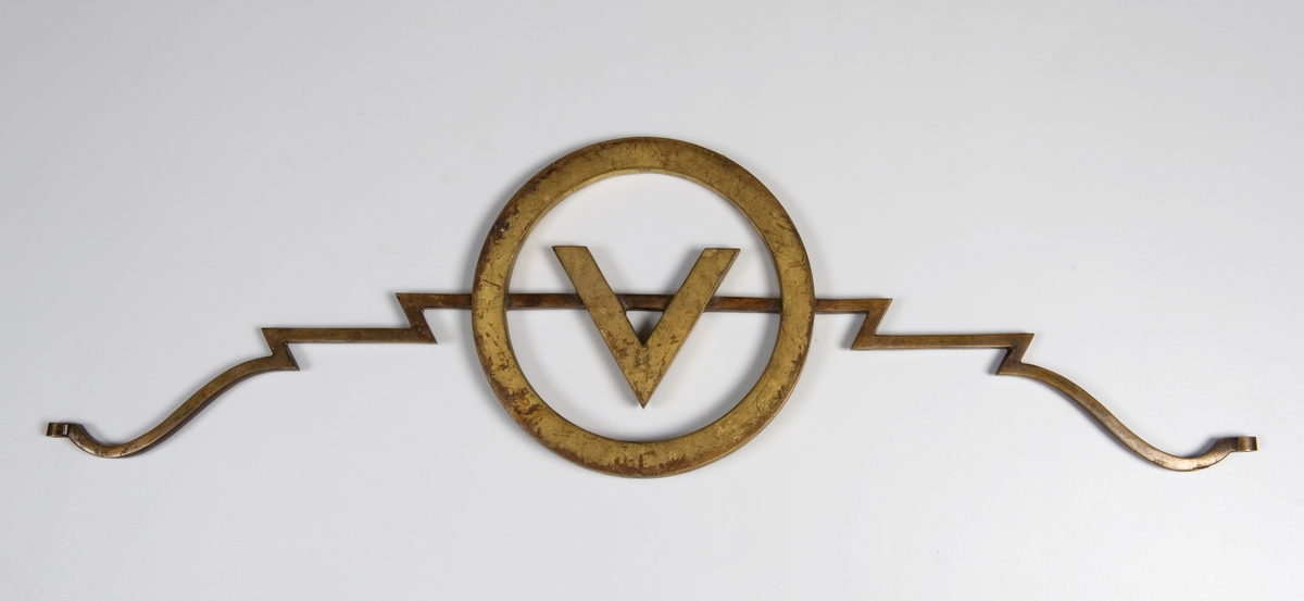 Toarmet gullfarget metallskilt med Vinmonopolets logo: V i sirkel.
