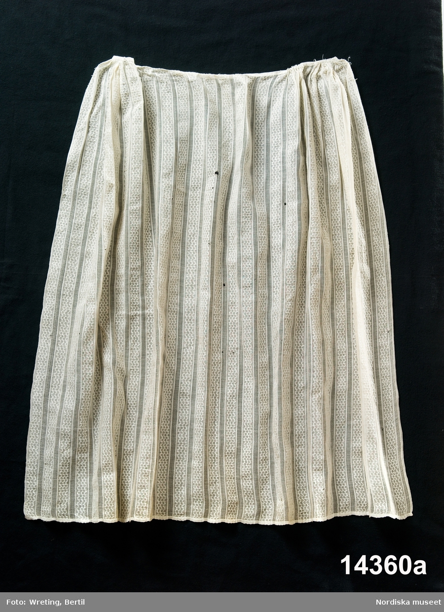 Förkläde av vit tunn bomullsväv, vävd med glesränder växlande med mönster i "lappet weaving" sicksackbårder i tjockare bomullsgarn. Rynkor vid sidorna, slätt på mitten mot smalt midjeband, knytband saknas.
Tyger som dessa masstillverkades i Skottlands tidiga bomullsindustri redan omkr. 1800. De användes  främst  till gardiner för fönster och som sänggardiner i borgerlig miljö. Blev mycket moderna i folkligt dräktskick omkr. 1830 och finns över stora delar av landet, användes mycket till brudförkläden och som konfirmandförkläden. Även avpassade kvadratiska kläden fanns i denna teknik och användes som huvudkläden.
Tygprovböcker med lappet weaving finns i museet i Paisley, Skottland.
/Berit Eldvik 2012-08-29