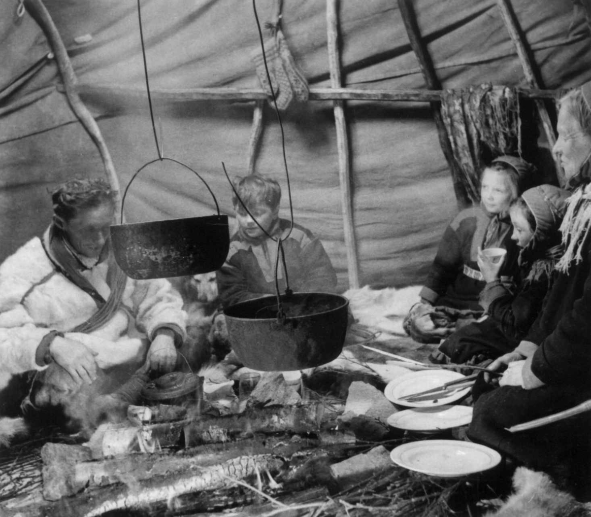 Familiene Sara og Eira sitter rundt ildstedet i teltet, kjøkkenutstyr er tatt frem. Karasjok 1958.