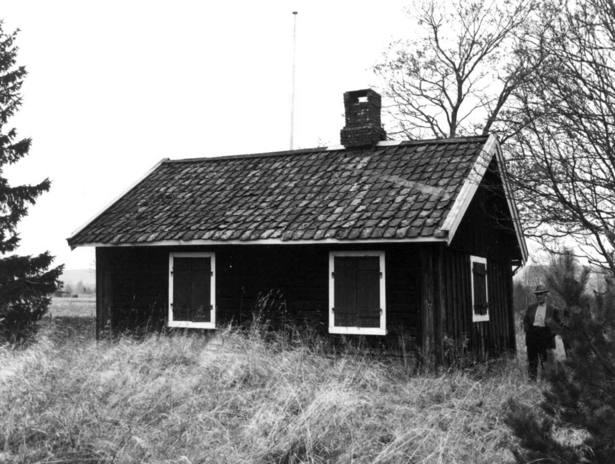 Enga husmannsplass, Asak, Skedsmo, Akershus 1964. Lite hus med lemmer for vinduene. Mann står ved hushjørne.
