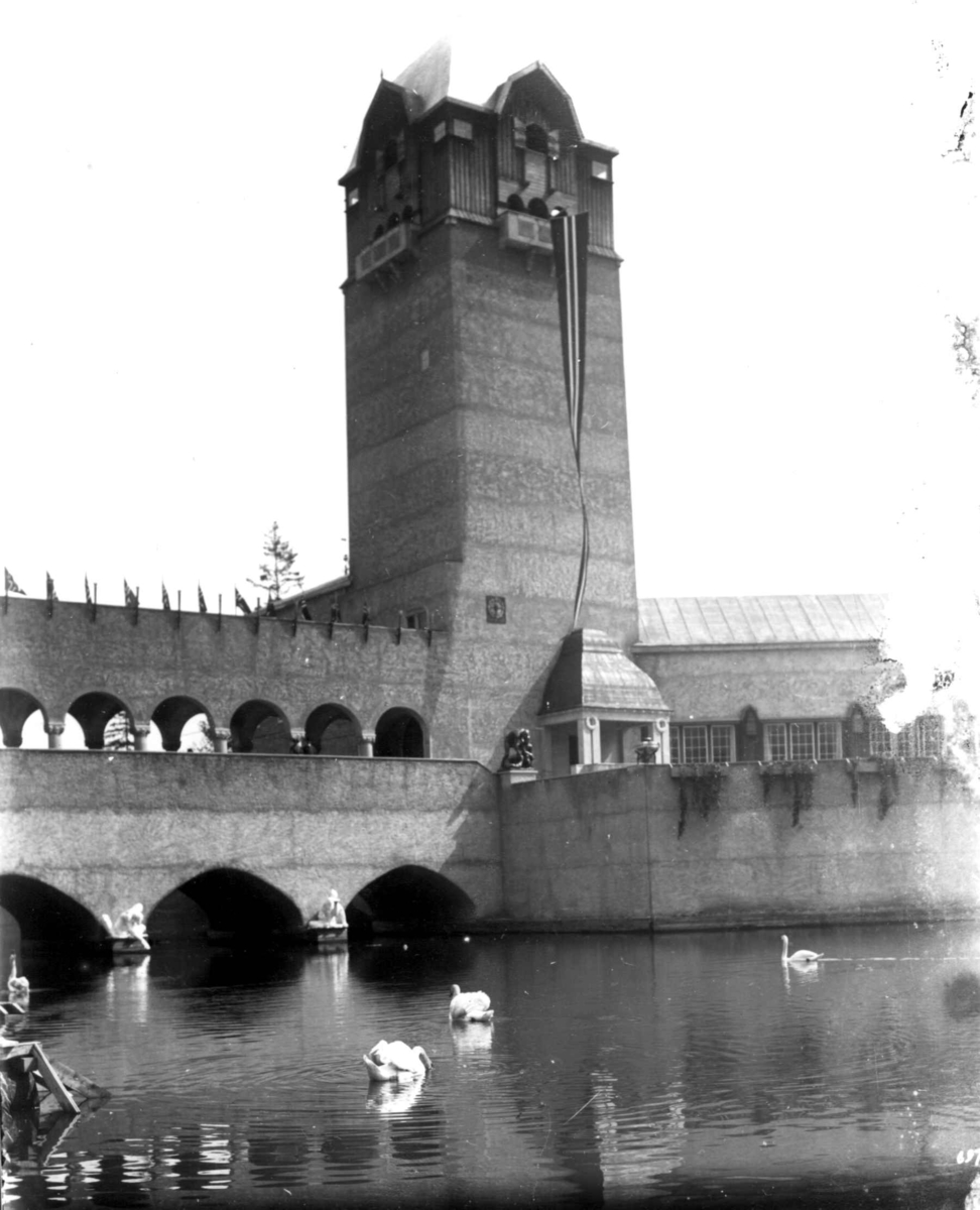 Jubileumsutstillingen på Frogner, Oslo, 1914.
Festbygningen.  Norsk vimpel og flagg. Vanndam med svømmede svaner i forgrunnen.
