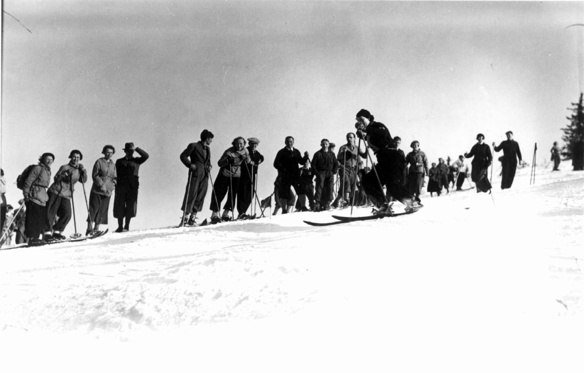 Slalåmrenn, Tryvannsåsen, Oslo. 1934. Kvinnelig skiløper i fart ned løypa. Tilskuere ses i bakgrunnen.