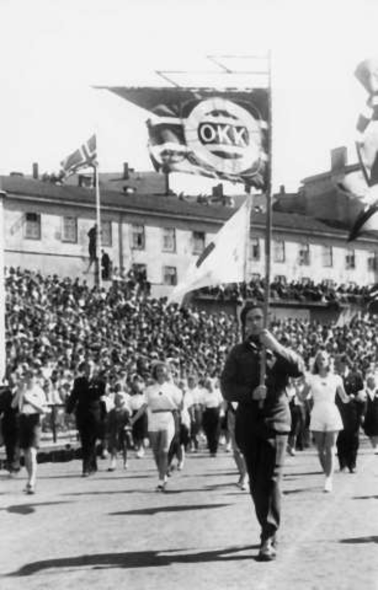 Fra Oslo under fredsdagene i 1945. Idrettens Dag på Bislett Stadion 3.juni. Det marsjeres på den oppmerkede banen. En ung mann i uniform bærer en fane merket OKK.