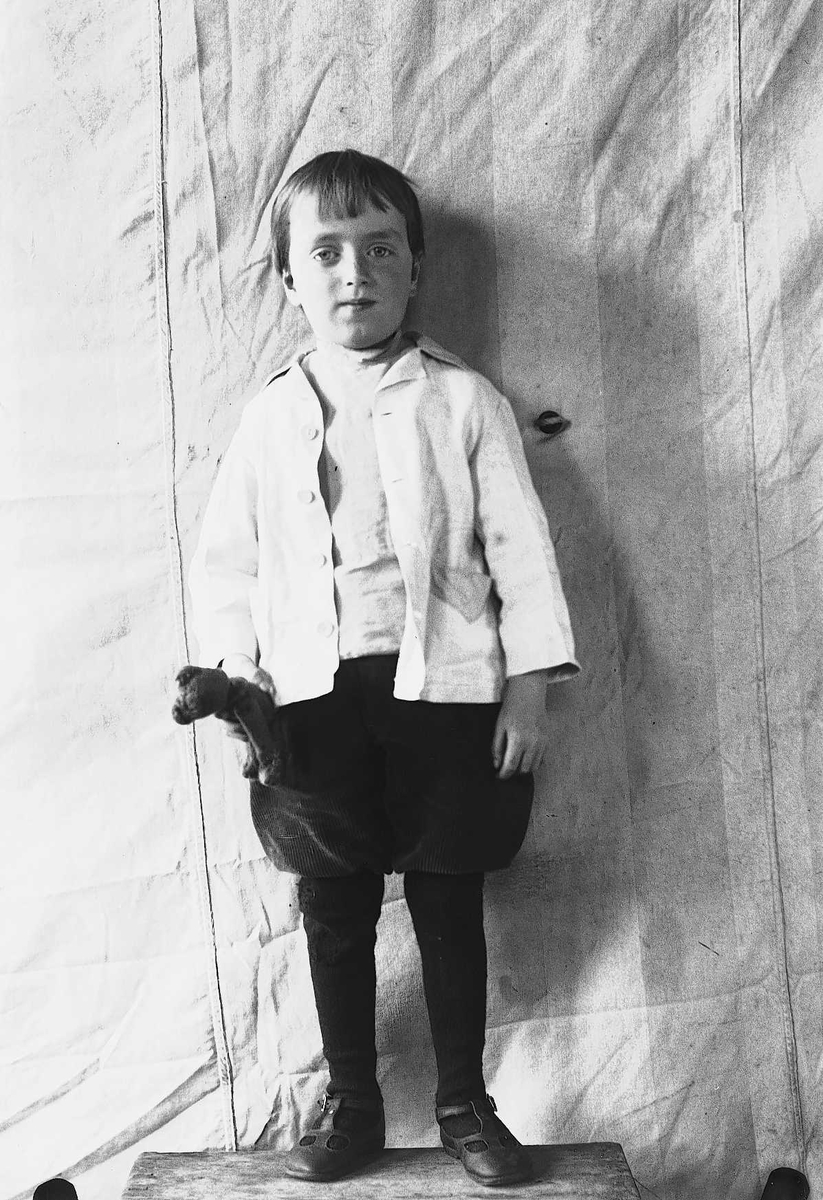 Portrett av gutt stående på krakk foran opphengt bakteppe, ukjent sted. Antatt en av barna i familen Axel Q. Wiborg.
