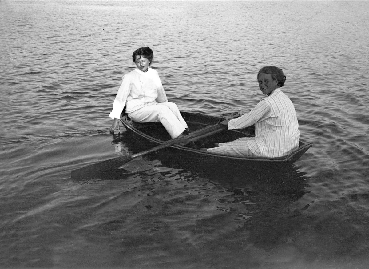 To kvinner sitter å ror i en robåt. Robsahm og Lund.