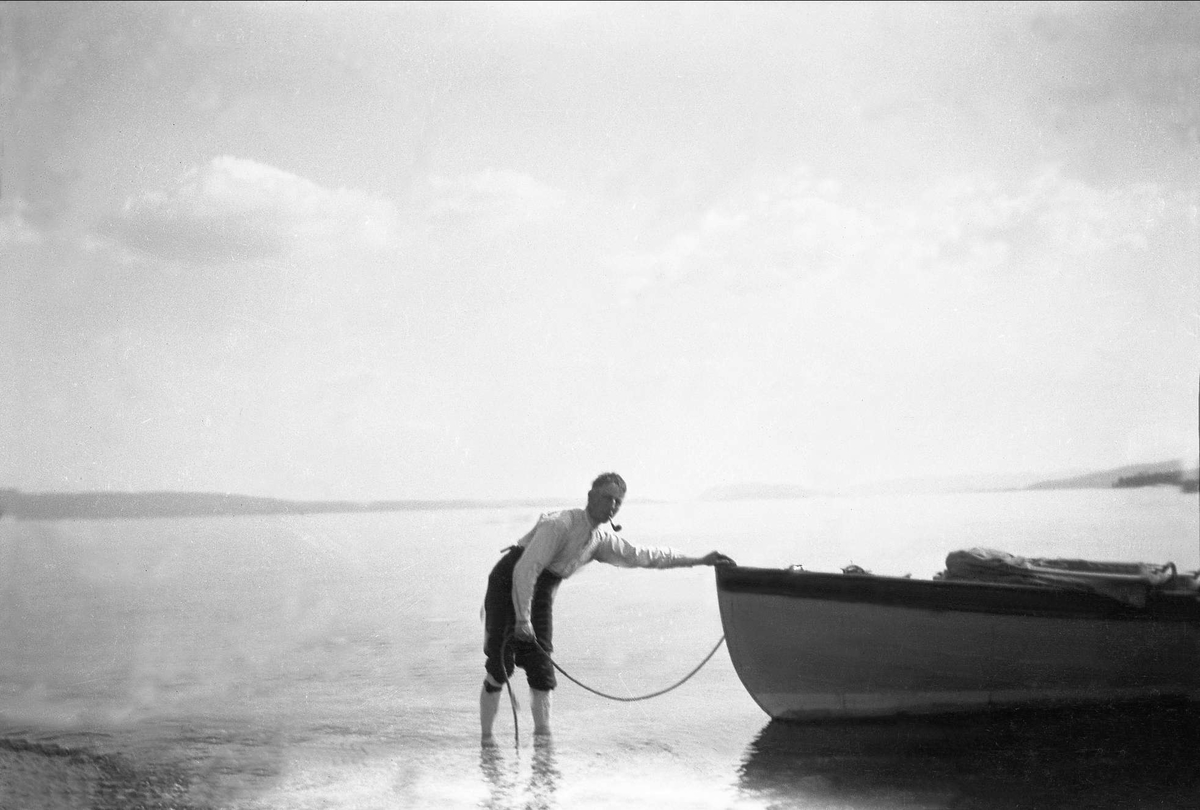 En mann med pipe i munnen står i sjøen og holder en båt med et tau. Robsahm og Lund.