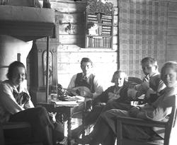 Fem unge mennesker samlet foran peisen på Lykkehytta. Fritjo