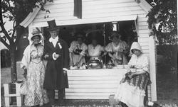 Postkort. Kvinner og en mann foran markedsbod med kaffekjele