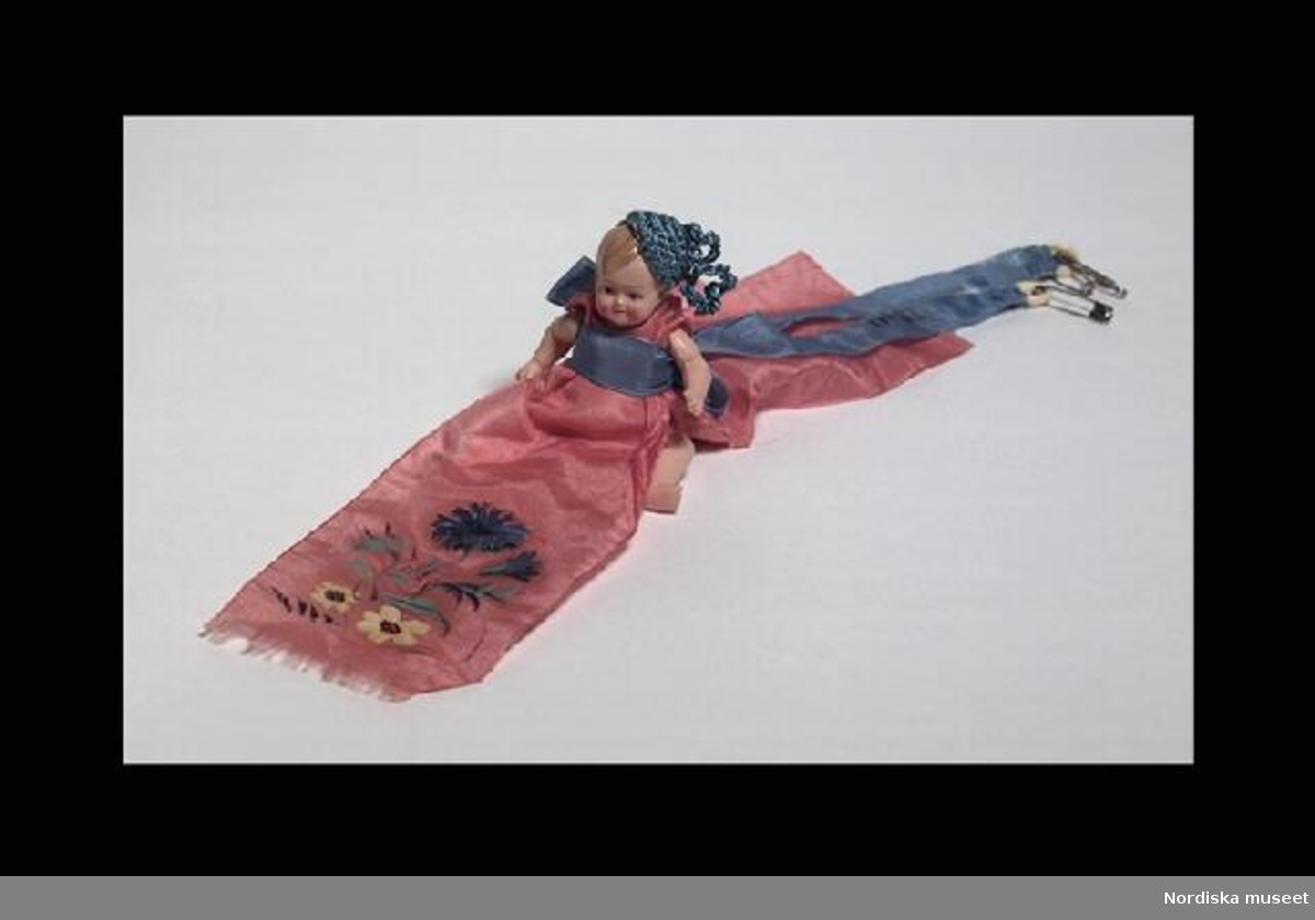 Inventering Sesam 1996-1999:
H  10 (cm)
Babydocka, använd som nåldyna, docka med ledade armar och ben av celluloid, målade ansiktsdrag och blont hår, iklädd  bärklänning av rosa sidenband med målad dekor av blåklint, skärp av blått sidenband i vars ändar hänger ringar av celluloid med 4 st säkerhetsnålar, under klänningen rosa tygstycke av flanell, använt som nåldyna (11 st knappnålar). Virkad mössa av blått garn.
Inpressat i dockans rygg: sköldpadda inom romb / GERMANY / 9 1/2" (=Rheinische Gummi und Celluloid Fabrik, Tyskland).
Ingår i samling föremål från Adolf Fredriks församlings arbetsstugor.
Anna Womack jan 1998
