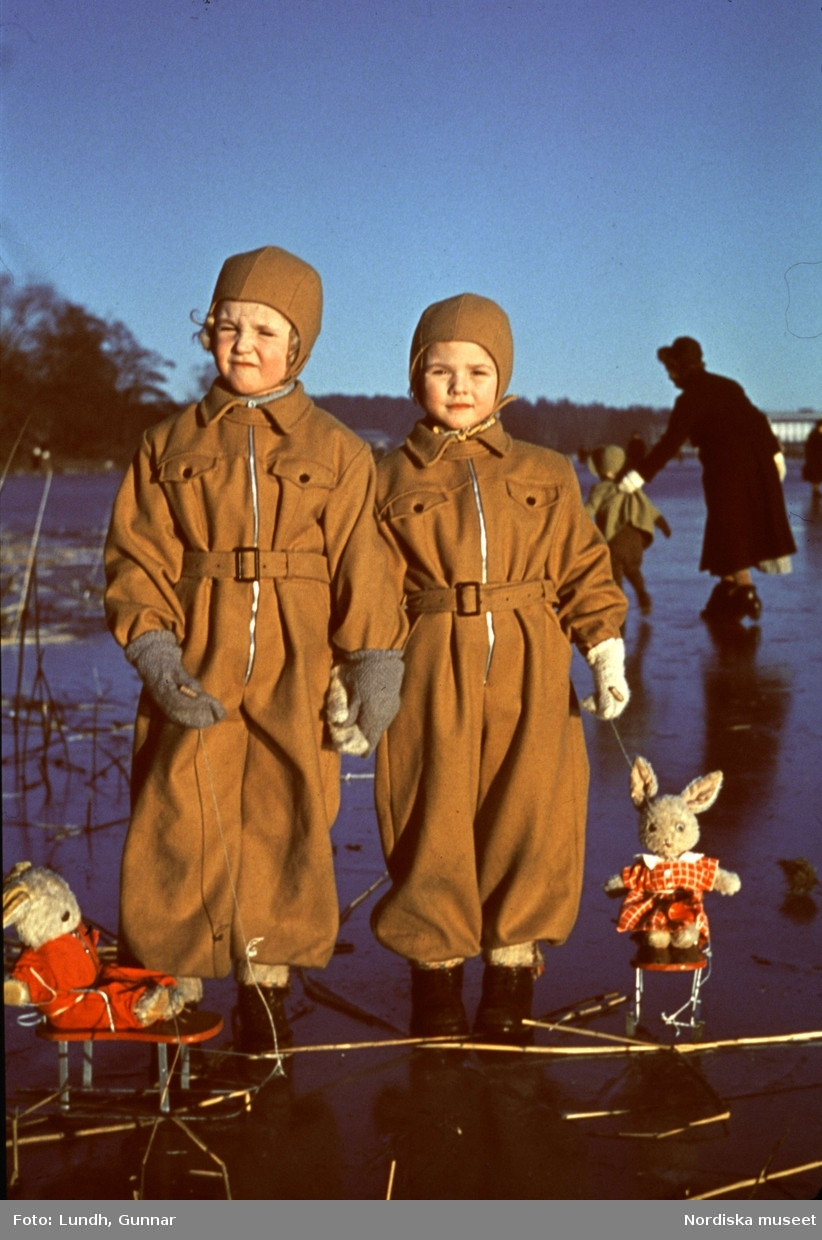 Barn med leksaker ute på is.Två flickor i overaller står på isen med varsin leksakskanin på små kälkar.
