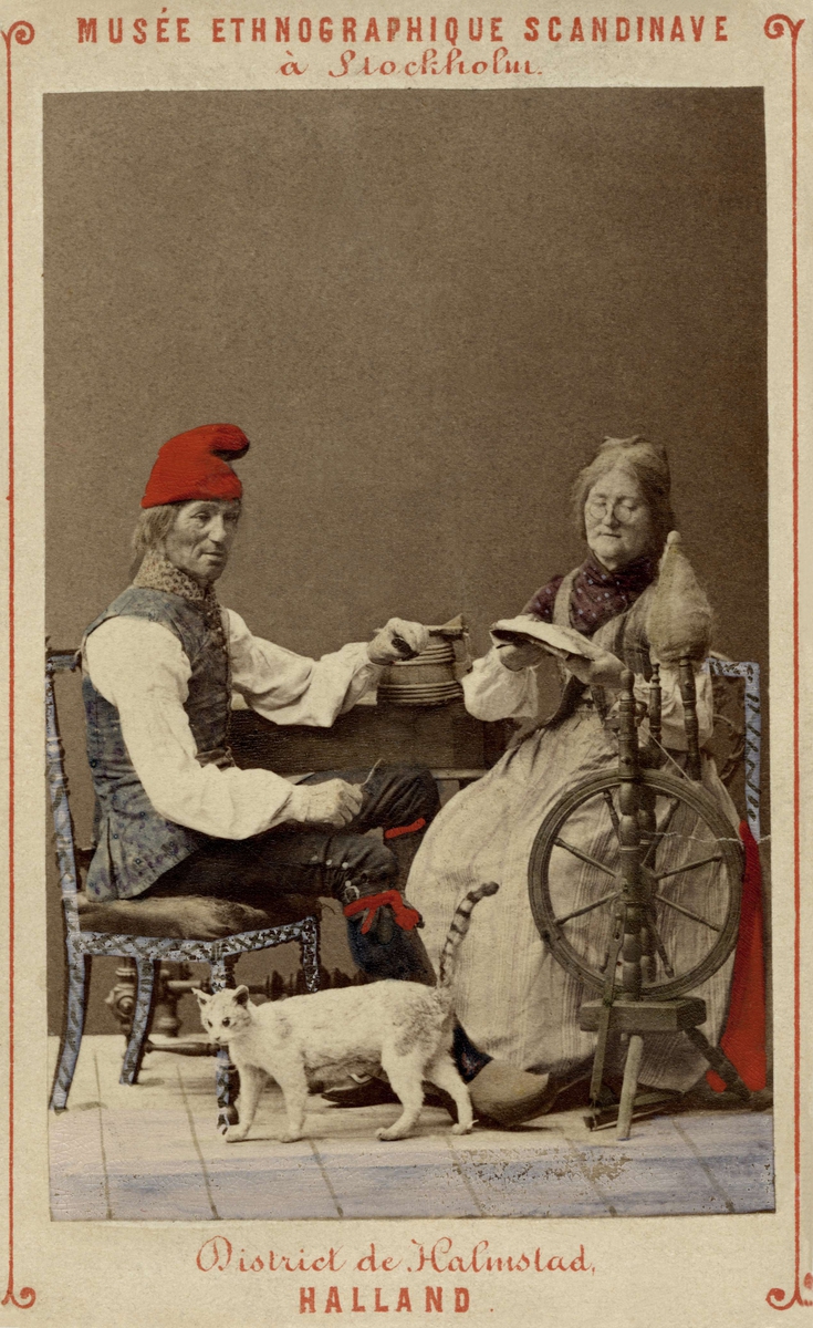 En man och kvinna i dräkter från Halmstad, Halland, sitter i ett kök, han snidar och hon spinner. Dräktdockor från Skandinavisk-Etnografiska samlingen i Stockholm. Handkolorerad.