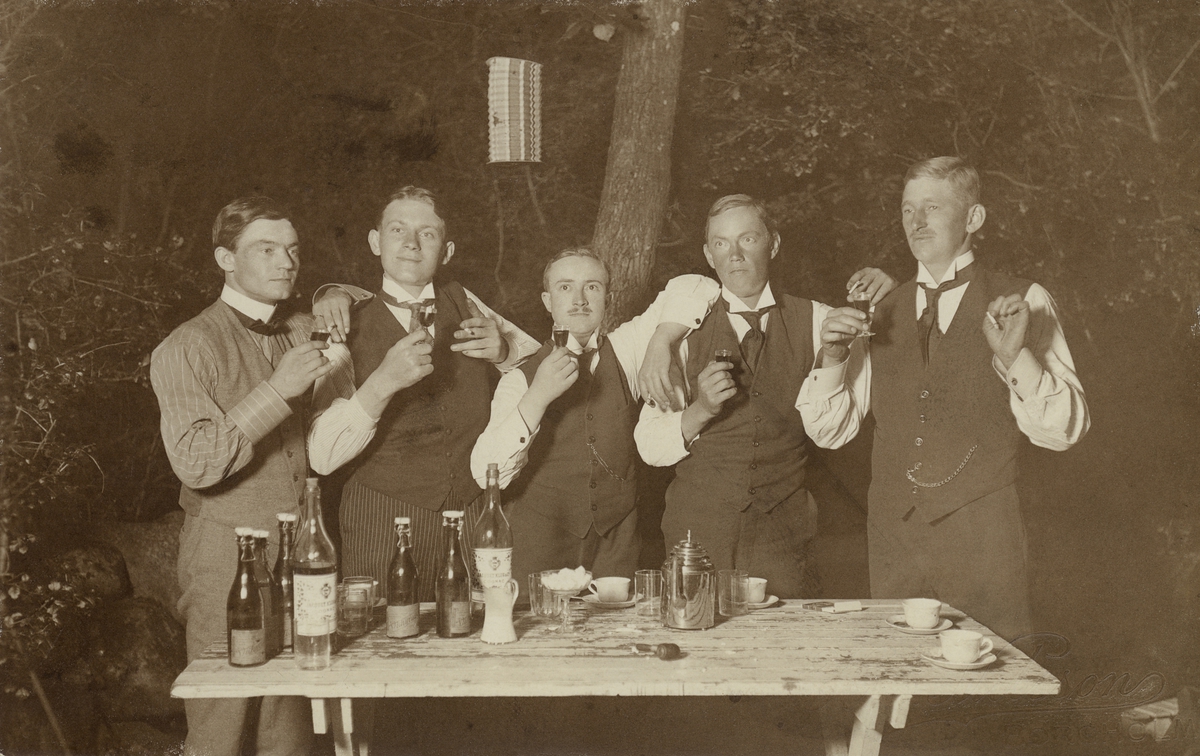 Fem män höjer sina snapsglas ute i en trädgård stående vid ett trädgårdsbord med kaffekoppar och flaskor.