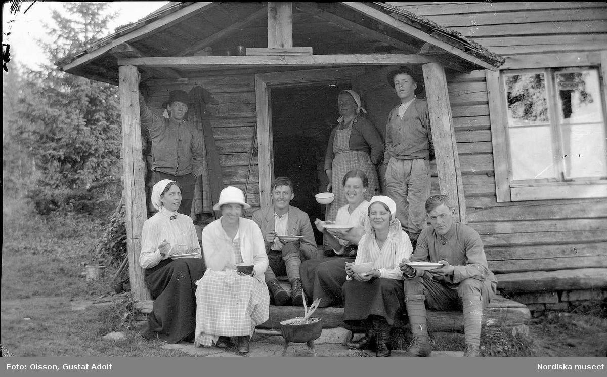 En grupp män och kvinnor sitter och äter på trappan till ett hus. 1900-talets första hälft.