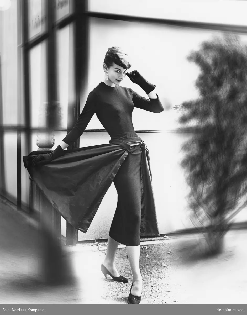 Kvinnlig modell poserar iklädd figurnära klänning med drapering och lösa flikar vid höften. Hon bär handskar, mockapumps och en Audrey Hepburn-inspirerad  frisyr.