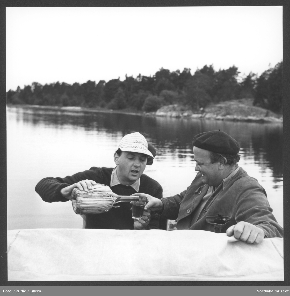 Författaren Stieg Trenter och fotografen KW Gullers på en båt i Gryts skärgård, Östergötland. Gullers häller upp ett glas vin ur en bastflaska.