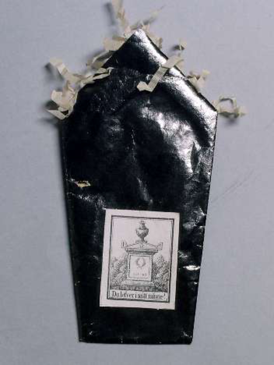 Begravningskaramell av svart glanspapper med vita fransar. Märke med gravvård och texten: "Du lefver i mitt minne".